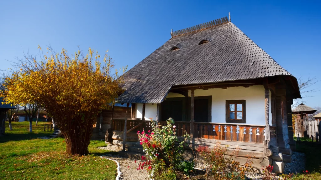 Casă tradițională cu pereți albi, acoperiș înalt din șindrilă și prispă din lemn cu motive decorative